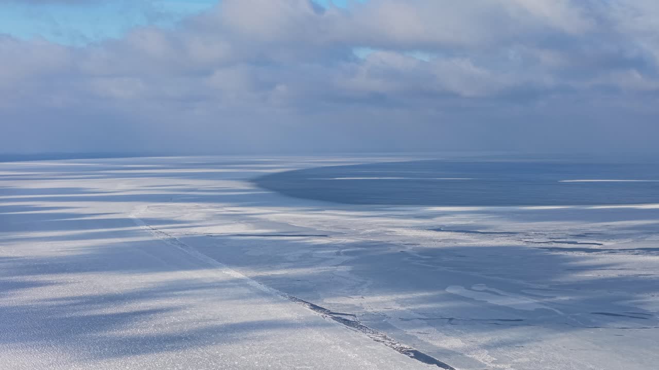 広大な凍結した湾：淡い冬の光の下、滑らかな氷の表面に映る、空撮された質感のある抽象的な雲の影。