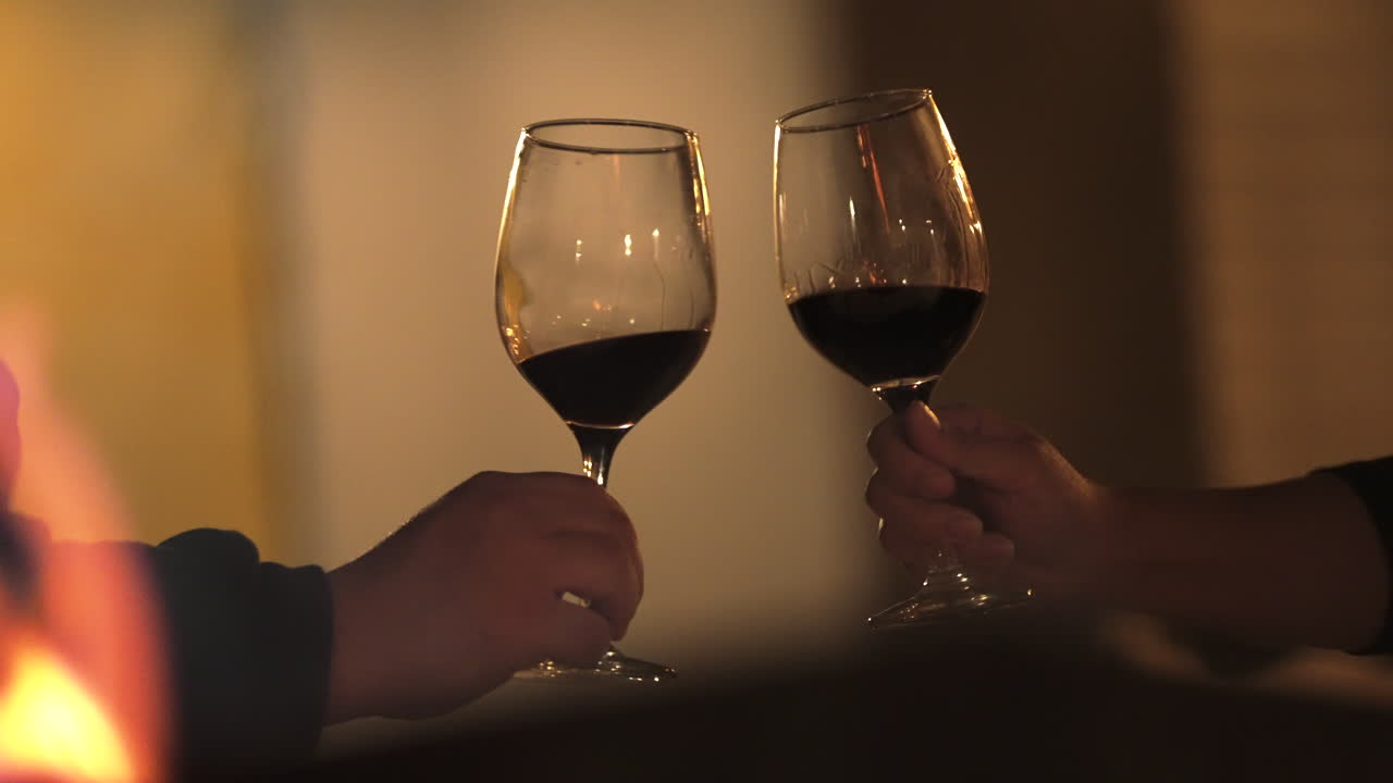 Primer plano de copas de vino tinto brindando cerca de una chimenea, manos de una pareja en una cita romántica