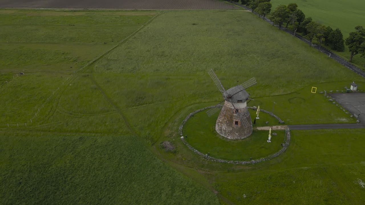 Scenic aerial view above Kuremaa traditional tower mill on overcast summer day. Drone moving backwards from historical windmill revealing dry stone walls and farmland, alley of trees by green field