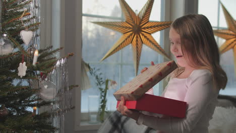 young girl in awe opening a christmas gift, indoor with a decorated tree and star ornament