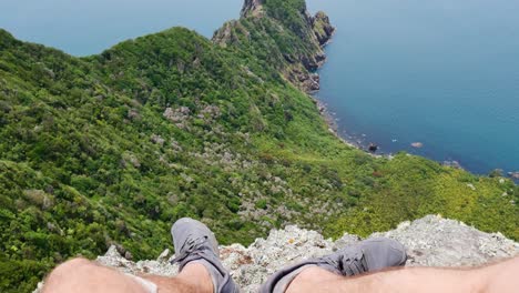 tilt up pov shot of male person in shorts enjoying beautiful ocean view and green vegetated mountains during sunny day from edge of summit - hiking at te whara track in new zealand