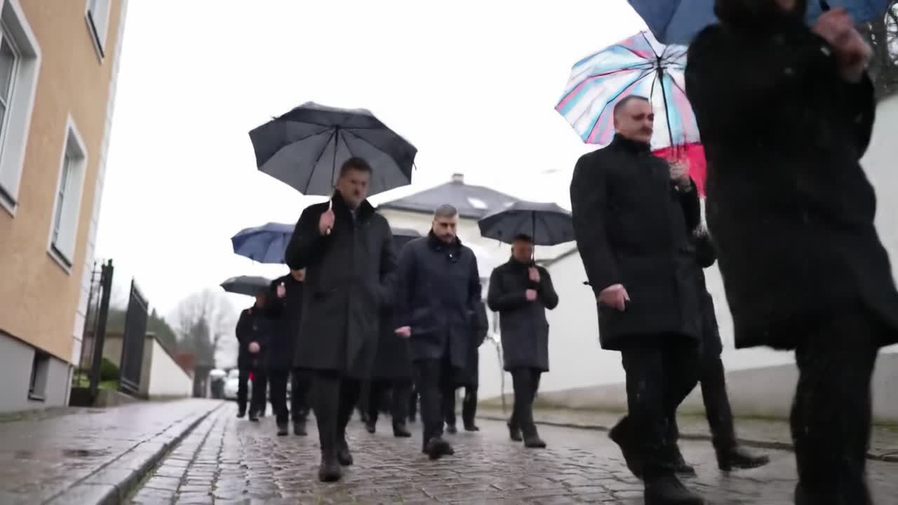 A solemn procession of individuals marching under black umbrellas, navigating a cobbled path on a rainy day, reflecting unity and remembrance in a muted atmosphere.