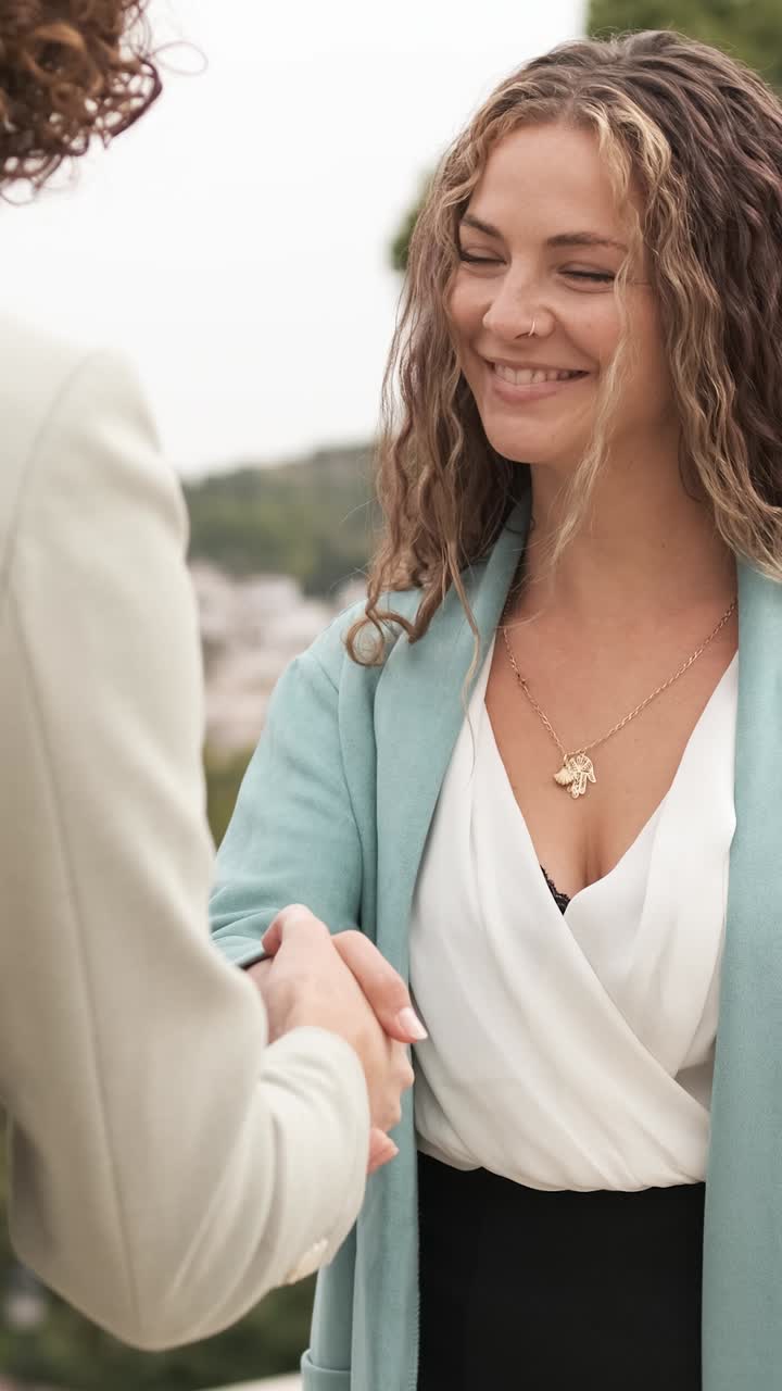 Businesswomen engaging in a handshake outdoors amidst a cityscape. Vertical