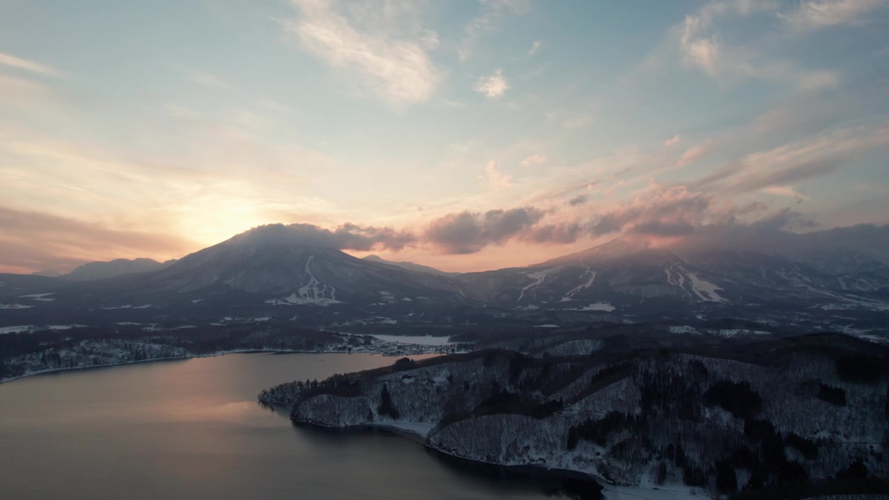 Penerbangan drone di Danau Nojiri (Nojiriko), Nagano, Jepang, menampilkan pegunungan di sekitarnya dan pemandangan matahari terbenam alami dengan salju yang mencair di sekitar pohon kering.