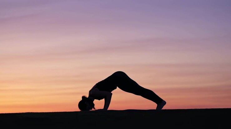 Silueta de perfil lateral de una mujer realizando una postura de yoga invertida en campo abierto al atardecer, con cielo pastel, horizonte bajo, contraluz tranquilo, equilibrio elegante y un ambiente de bienestar al aire libre concentrado.