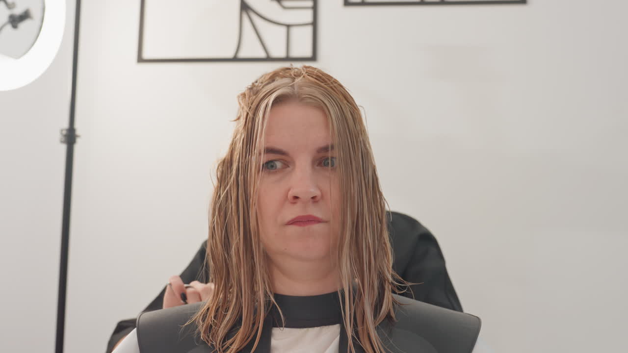 Donna caucasica seduta su una poltrona in un salone di bellezza che riceve un trattamento di treccine sotto la luce di uno studio, parrucchiera che sistema le micro trecce con dita curate, espressione calma e concentrata, interni minimalisti bianchi, primo piano
