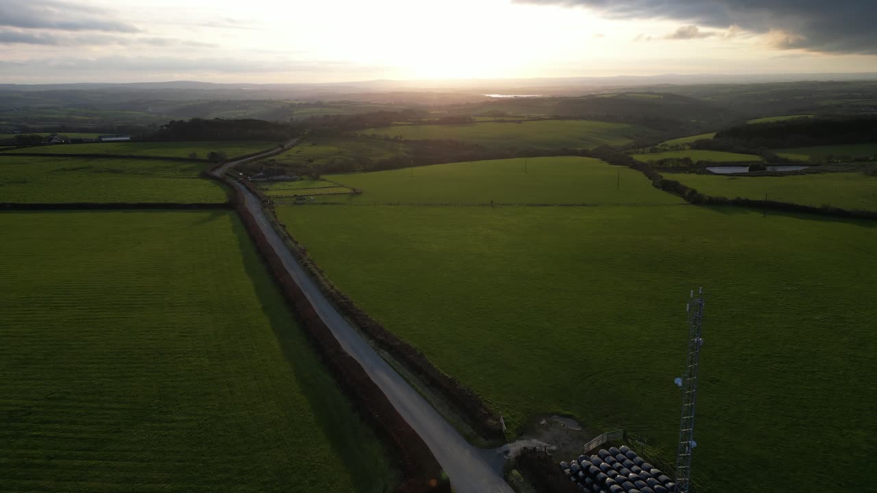 Stunning drone shot of a winding rural road surrounded by green fields during sunset. The warm light creates long shadows, highlighting the tranquil countryside and distant hills.
