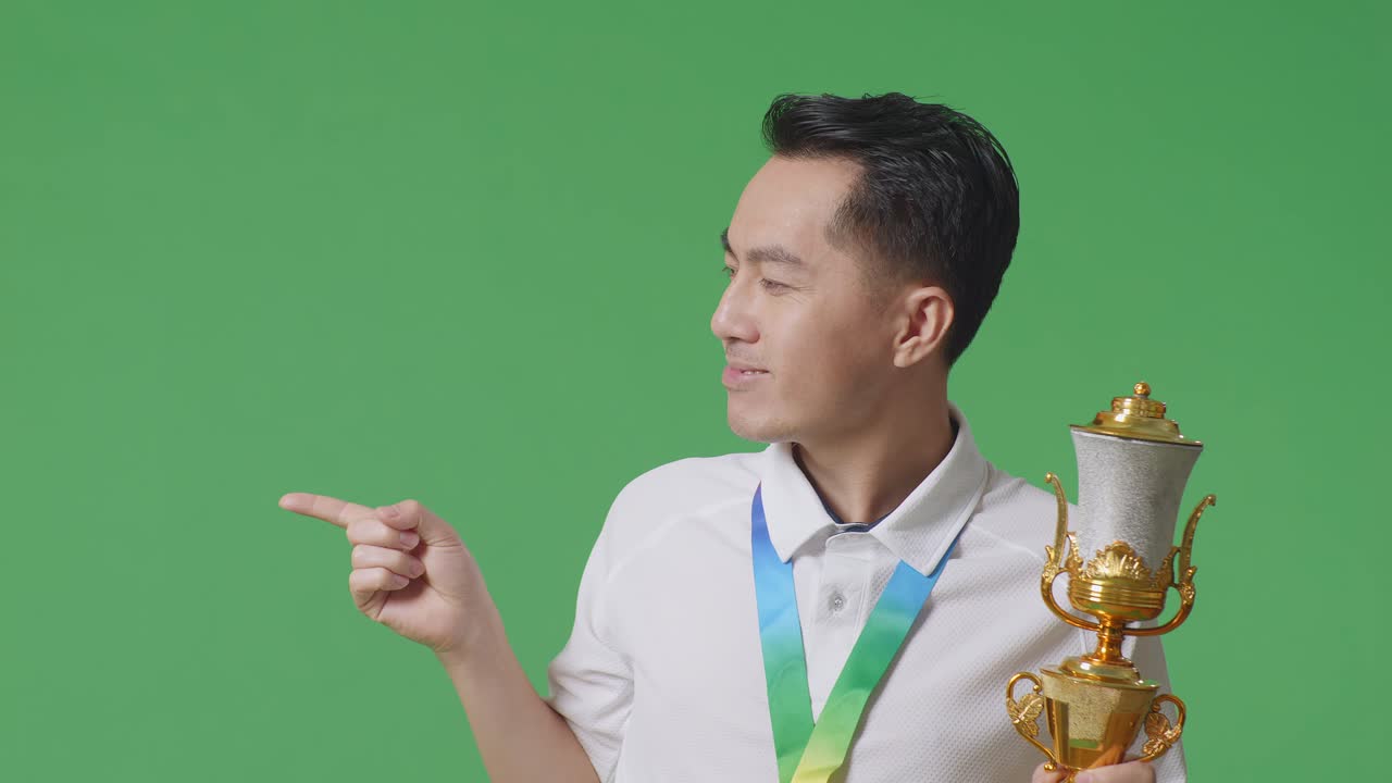금메달을  아시아인 남자가 손에 금 트로피를 들고 미소 짓고 스튜디오의 녹색 화면 배경에 을 가리키고 있습니다.