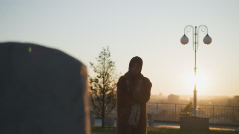 a grief-stricken girl stands in front of a stone monument in a park during sunset, her hands folded in her arms, wearing a coat and scarf. the warm, golden light enhances the sorrowful mood