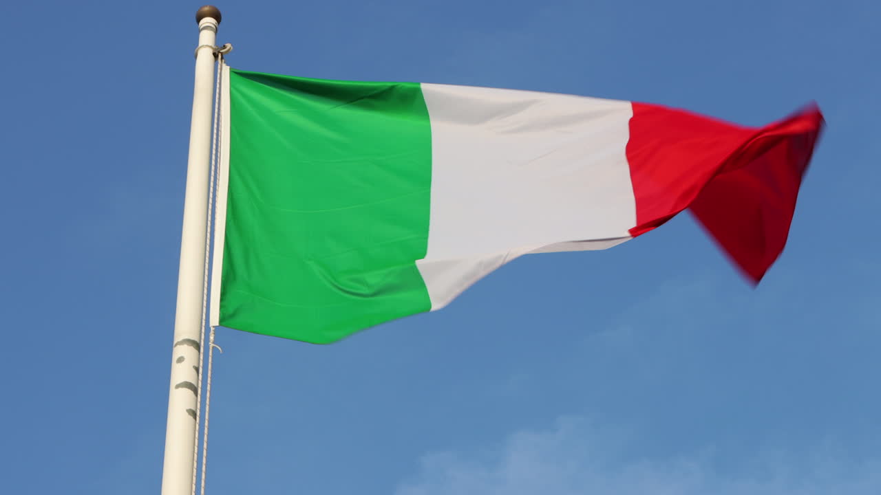 Inquadratura dal basso della bandiera italiana che sventola al vento contro un cielo azzurro limpido.