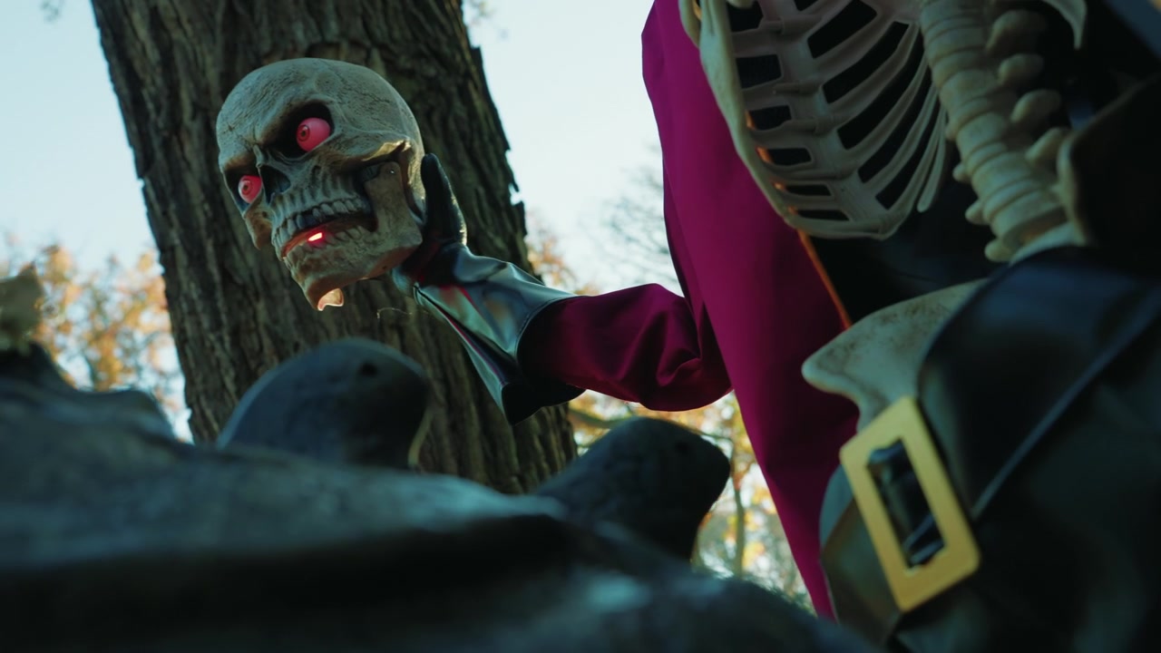 Nærbilde av et skjelett som holder en glødende hodeskalle med røde øyne, kledd i en rød kappe og belte, plassert mot et tre i en utendørs Halloween-scene.