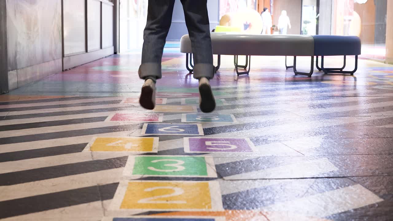 An adult in casual shoes and jeans jumps along a colorful hopscotch path inside a brightly lit indoor playground with benches and striped flooring