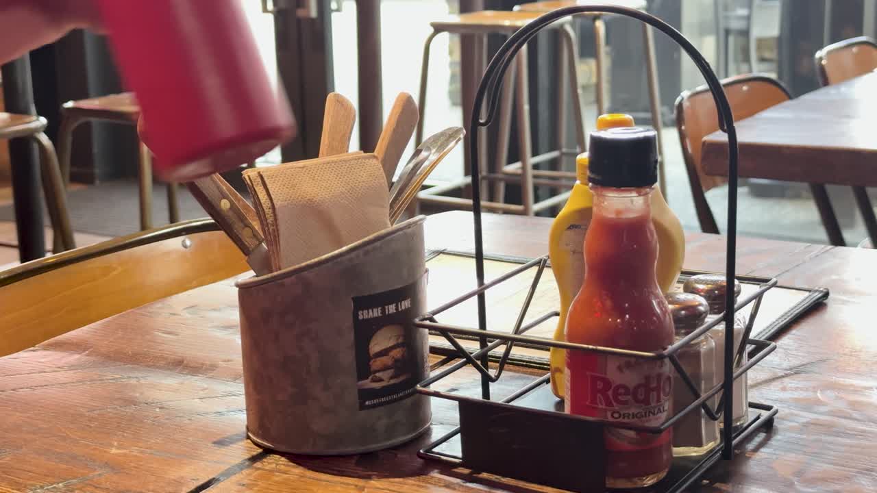 A hand reaches for a ketchup bottle on a cafe table, surrounded by condiments and utensils, in warm lighting