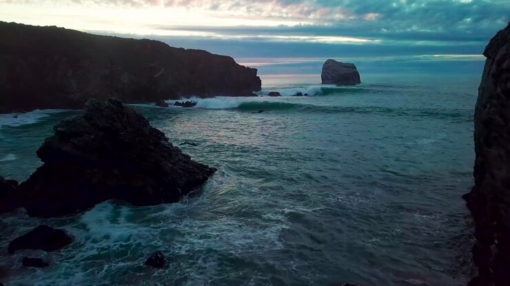 Volando vicino a enormi rocce oceaniche, accanto alle onde che si infrangono, verso il sole che tramonta a Sand Dollar Beach a Big Sur, California.