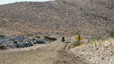 Moto-rider-drives-arid-hillside-gravel-road-around-distant-curve