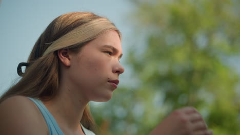 thoughtful young woman outdoors with sunlight softly illuminating her face, adjusting her hair back pinned with black pin, wearing cyan top, under blurred sky, with blurred view of green tree