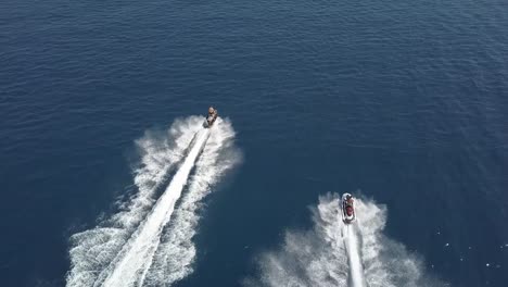 Tiro-De-Seguimiento-De-Dos-Personas-En-Jet-Ski-Montando-De-Lado-A-Lado-En-El-Mar-Azul-Abierto,-Chipre