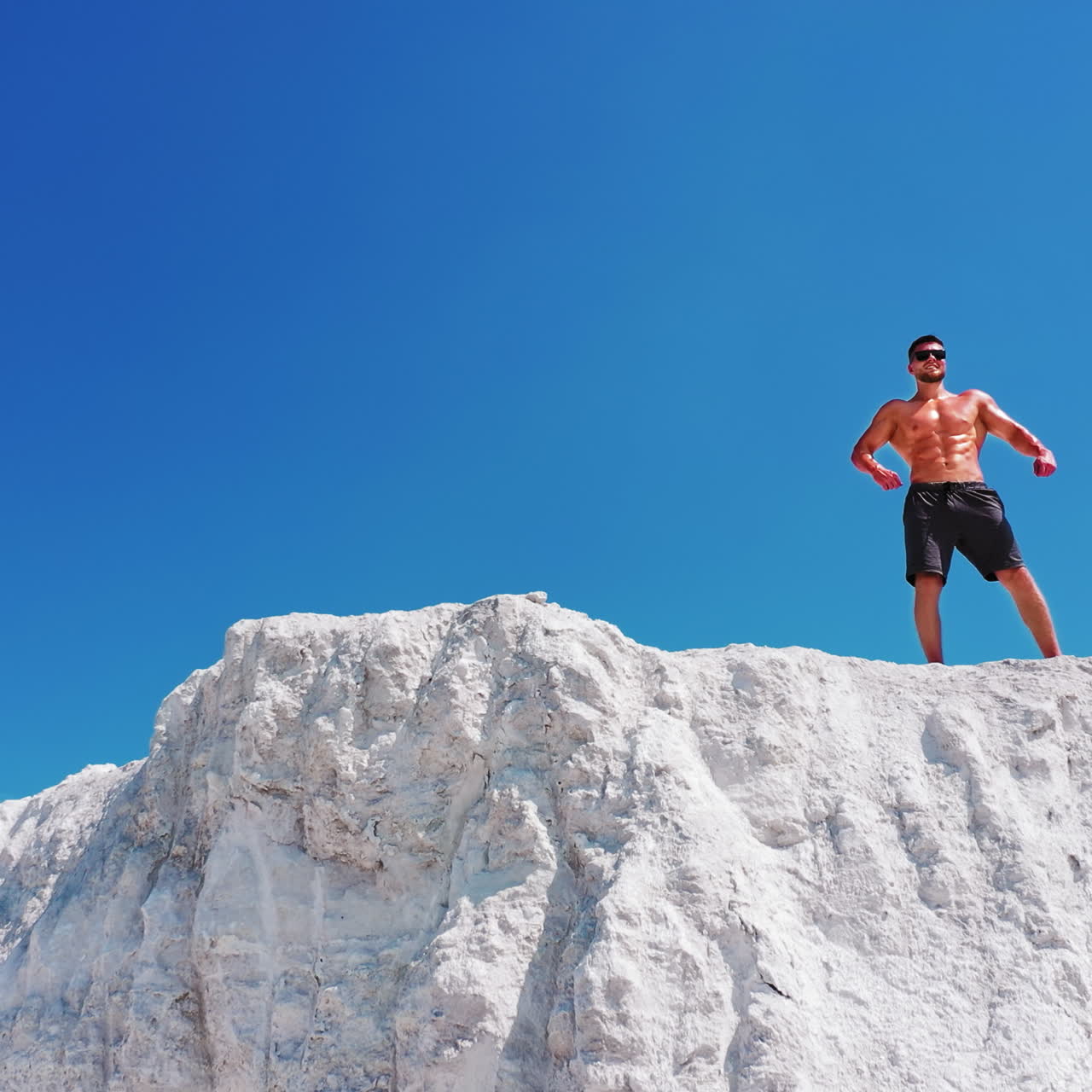Handsome man in shorts with muscular body standing on the white hill under blue sky. Bodybuilder without shirt posing on the top of the mountain. Drone view.