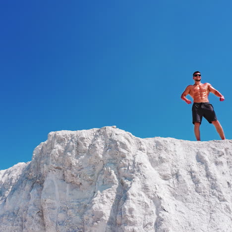 handsome man in shorts with muscular body standing on the white hill under blue sky. bodybuilder without shirt posing on the top of the mountain. drone view.