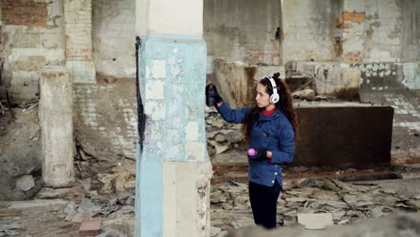 serious pretty girl is busy painting graffiti on old dirty pillar in empty industrial building using spray paint while listening to music with headphones.