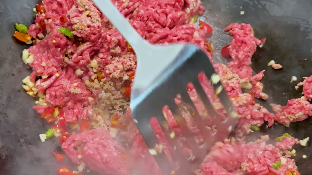 Raw minced meat is stirred with chopped vegetables and spices in a hot wok, using a spatula. Bright lighting, close-up, dynamic movement
