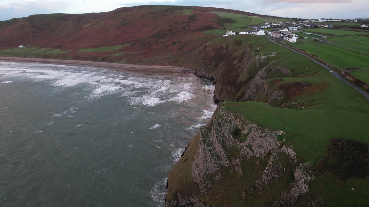 Aerial view of Rhossili Bay with green cliffs, red hills and sandy beach revealed as a drone slides left toward the ocean on the Wales coast