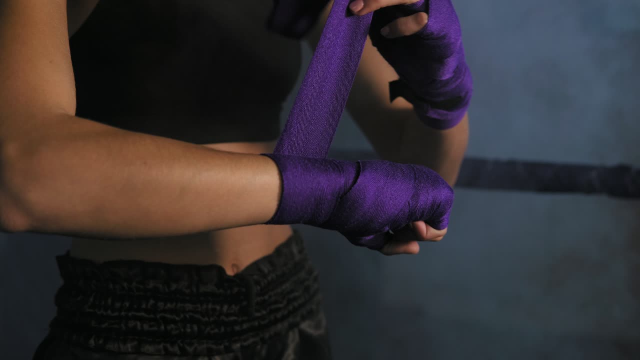 vista en primer plano de manos femeninas siendo envueltas para el boxeo en una habitación oscura con humo. joven mujer en forma envolviendo las manos con vendaje de boxeo púrpura. filmado en 4k