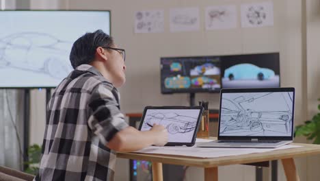 vista lateral de un hombre asiático con dolor de espalda mientras dibuja un nuevo concepto de diseño de automóvil en una tableta en el estudio con tv y computadoras muestra modelo de automóvil eléctrico 3d