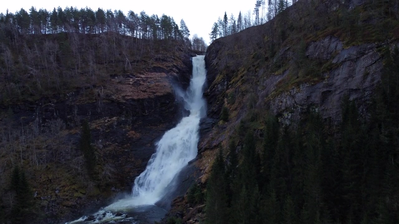 Stigende luftfoto av en foss som kaskader ned en klippe og ned i et basseng, omgitt av eviggrønne trær i en fjellrik norsk skog.