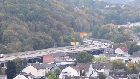 wide shot of traffic on german autobahn highway with many cars and trucks beside green mountain and housing area