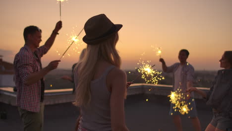 The-young-women-in-the-hat-beautifully-moves-her-hands-in-a-dance-with-big-bengal-light.-She-is-on-the-party-with-her-friends-on-the-roof.