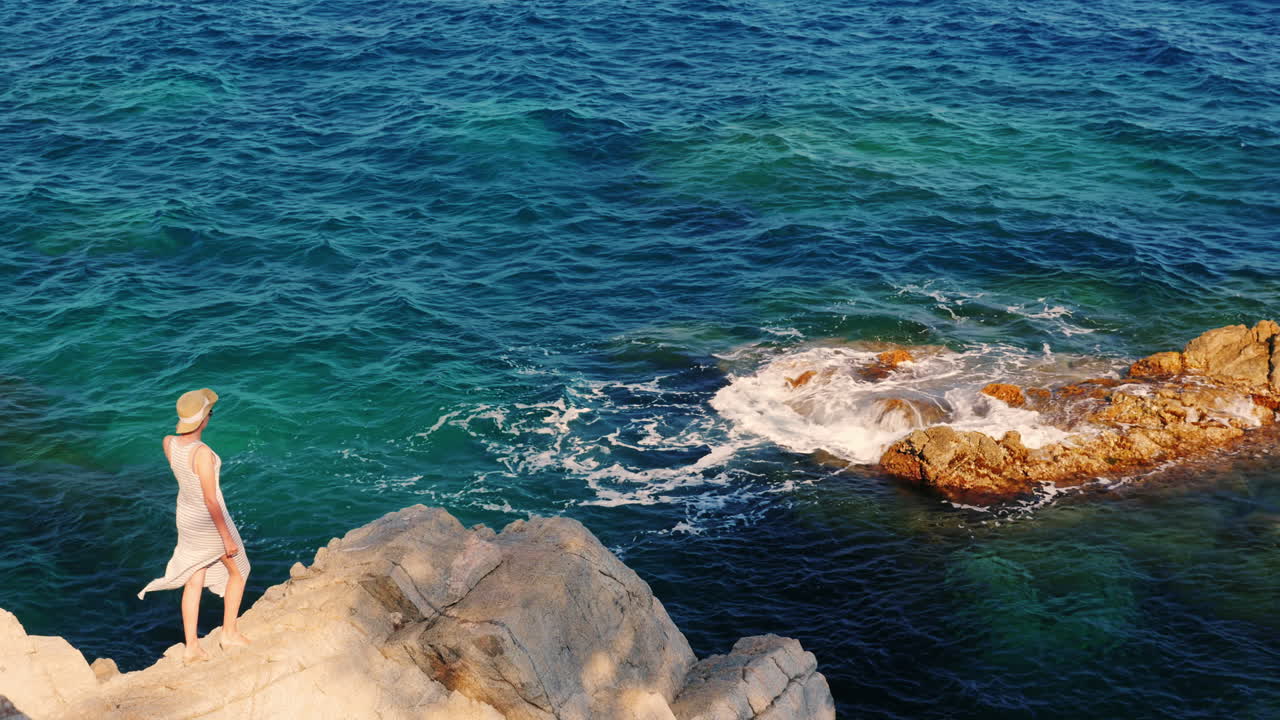 женщина в светлом платье и шляпе стоит на скале на фоне моря ветер дует неторопливо на се