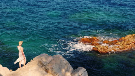 una mujer con un vestido ligero y un sombrero de pie sobre una roca contra el mar el viento sopla ocio en se