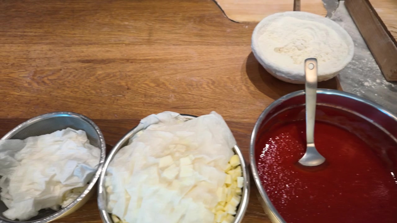 La telecamera dall'alto scorre su ciotole di salsa di pomodoro, mozzarella e farina su un bancone di legno.