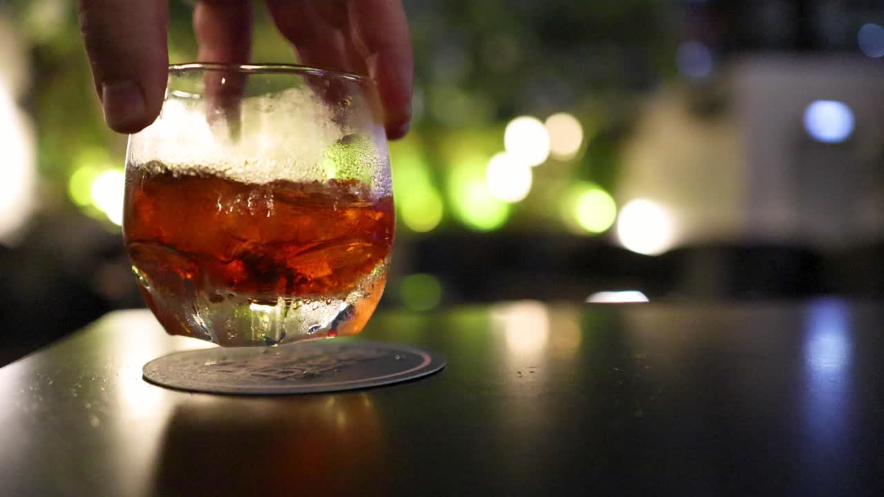 A hand gently swirls an amber cocktail in a glass on a dark bar table, with warm ambient lighting and soft background bokeh in a Bangkok nightlife setting