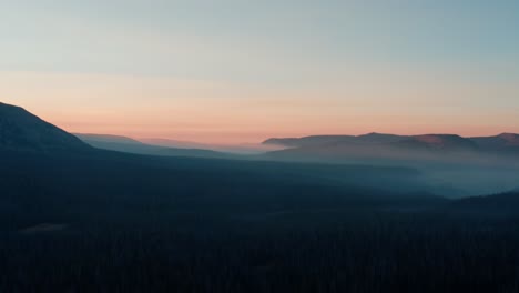 Hermosa-Toma-Aérea-De-Drones-En-Ascenso-Del-Impresionante-Bosque-Nacional-Salvaje-De-Uinta-Wasatch-Cache-En-Utah-Con-Grandes-Pinos-Debajo-E-Impresionantes-Montañas-Cubiertas-De-Niebla-Durante-Un-Amanecer-De-Verano