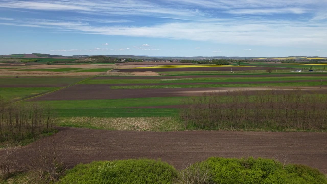 Eine weite Luftaufnahme fängt ein lebendiges Flickenteppich aus landwirtschaftlichen Feldern unter einem klaren blauen Himmel ein und zeigt die Schönheit der ländlichen europäischen Landschaft im Frühling.