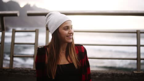 Smiling-Girl-Sitting-With-Longboard-At-The-Border-At-Coastline-Asphalt-Road-On-Background-Hills-And-Foggy-Ocean