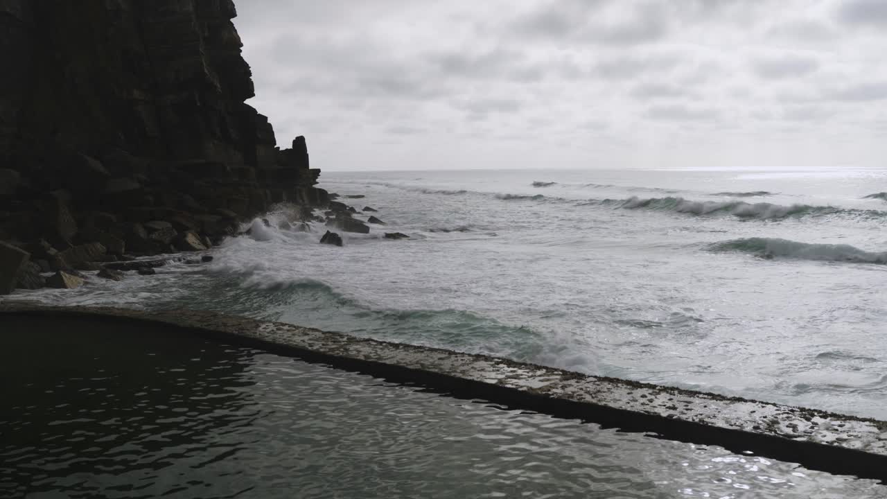 Static view of ocean waves hitting rocky cliffs near a natural tidal pool under a moody, overcast sky