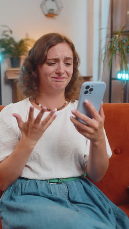 upset displeased caucasian woman using smartphone reading bad news sitting on couch in living room