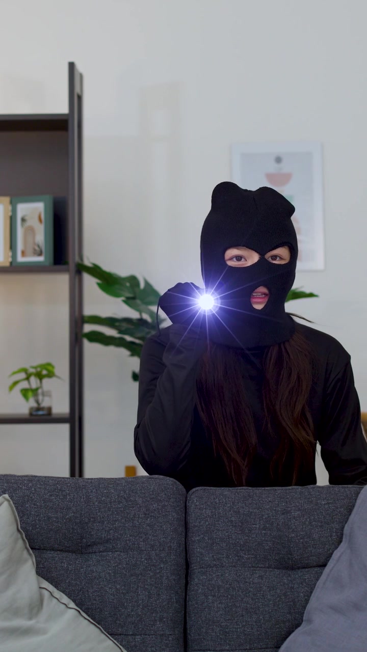 A masked intruder searches a living room at night using a flashlight behind a sofa