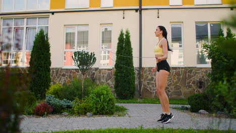 A-young-beautiful-girl-jumps-on-a-skipping-rope-in-a-Park-in-slow-motion-in-sports-clothes.-Play-sports-against-the-background-of-houses