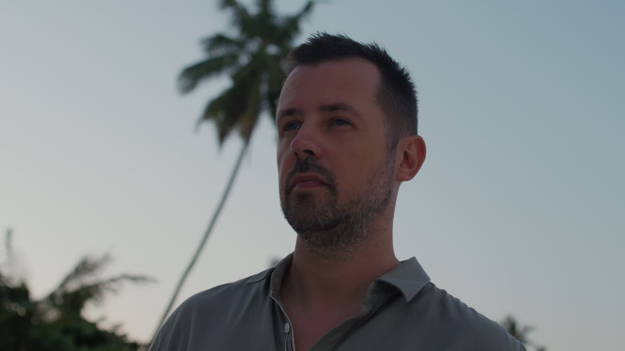 de cerca en fresco, confiado, hombre aspirante quitando frescas gafas de sol caminando a lo largo de la playa tropical en la puesta de sol