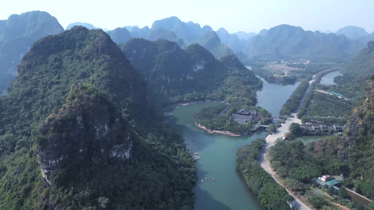 Drone footage of a large temple complex nestled beside a calm river, surrounded by dramatic limestone mountains and dense tropical forest in Southeast Asia.