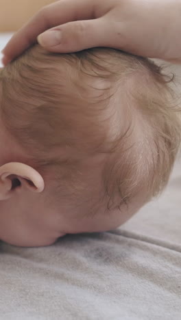 restless and adorable infant son lies on stomach on changing table and mother pets baby blond head extreme close view