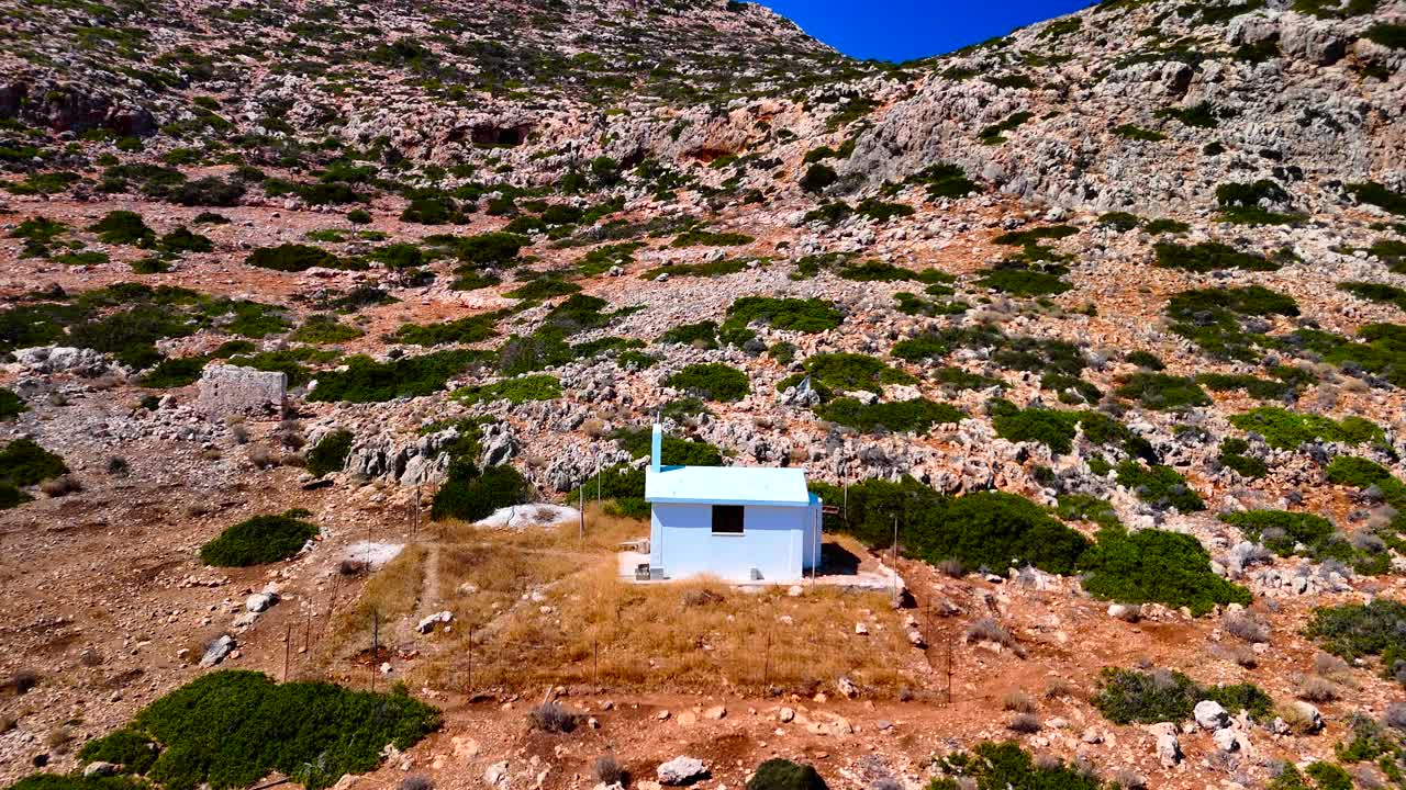 A small blue house sits alone in a rocky terrain, surrounded by sparse vegetation. The clearing brightens under midday sun, showcasing the beauty and isolation of this coastal location