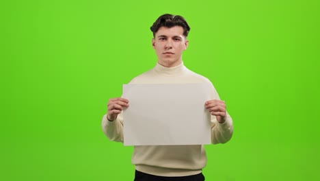 el hombre está de pie y mirando a la cámara. está levantando un papel blanco. está de pie sobre un fondo verde. pantalla verde. 4k.