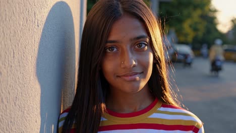 young woman smiles softly while standing against a sunlit wall during the golden hour. shadows and soft lighting create a warm atmosphere on a peaceful street