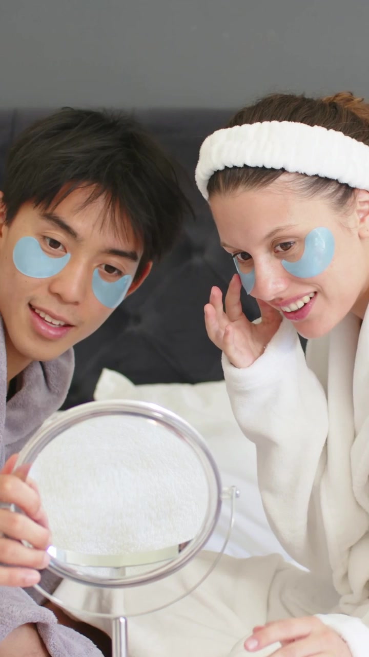 Vertical video: Holding mirror, woman in robe checking blue eye patches on bed, partner joining