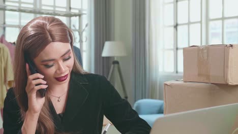 Young Asian Transgender Woman Business Owner With Boxes On The Table Happy Online Sales Job, Get An Order From Customers, Talking On Phone And Writing On Notebook
